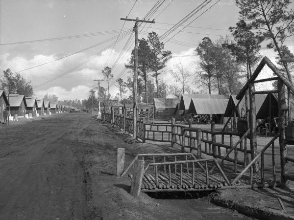 A view down Regimental Street, a dirt road at Camp Logan, with a bridge leading from the street to buildings on the right.  Small buildings also line the left side of the road, and a telephone pole stands near a fence.