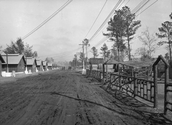 A view of two automobiles driving down Regimental Street, a dirt road at Camp Logan, where a man is standing by one of many buildings on the left.  The road is bordered by a fence on the right, and a telephone pole stands in the distance.