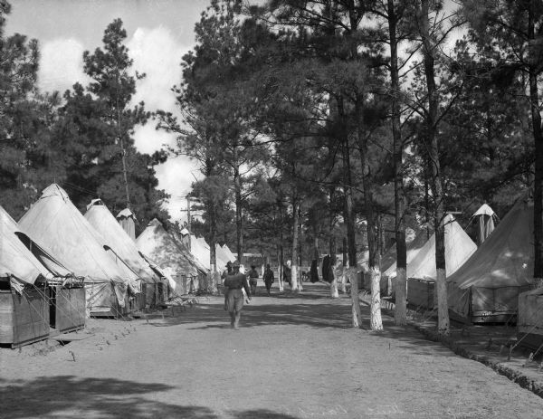 Men in uniform walk down a dirt road with tents on either side, surrounded by trees.