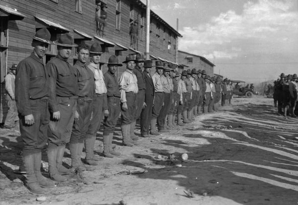 A row of soldiers at Camp Upton, created in 1917.  Men observe the formation of soldiers from the windows behind them and an automobile can be seen in the background.