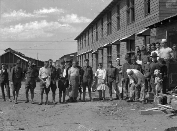 An informal portrait of soldiers standing near their barracks.  On the right, a soldier holds a dog.  The barracks and other military buildings stand in the background.