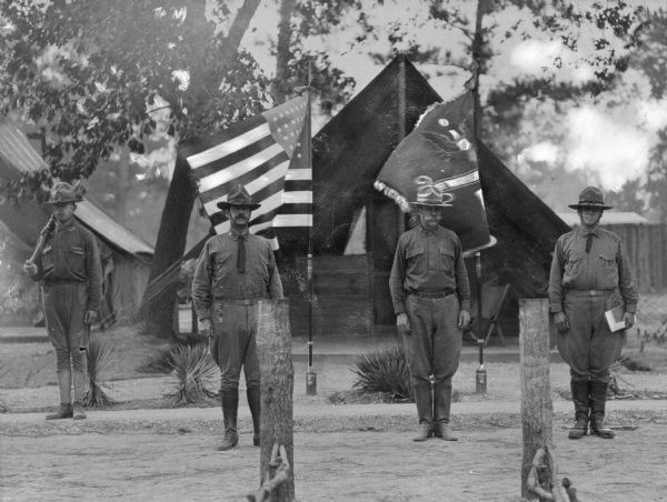 Four solders stand before two flags and their tents. The soldier on the left holds a rifle.