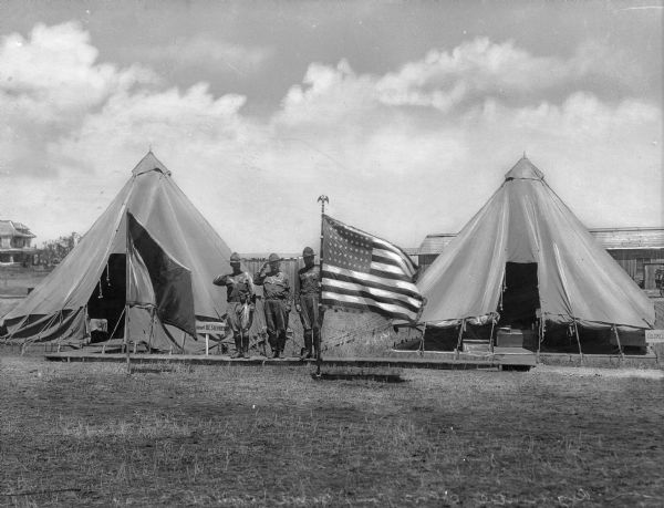 Army soldiers stand at attention near the United States flag and their tents at Camp Bowie.  Military buildings are visible in the background.