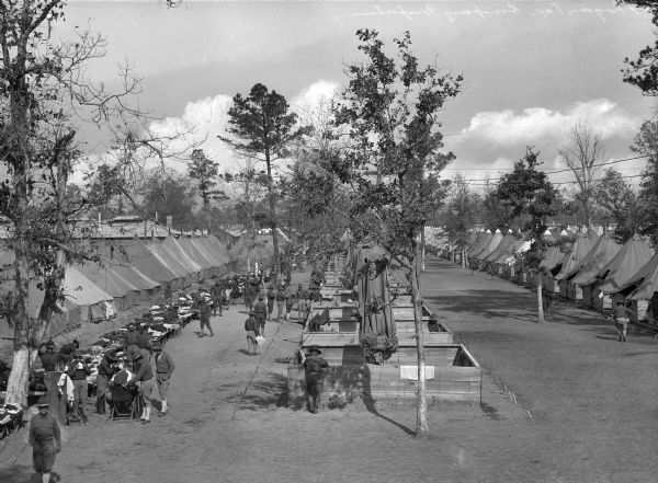 Elevated view of a company inspection with the tops of tents drawn back. Cots and equipment are lined up outside the tents and men walk on either side of the dirt road.