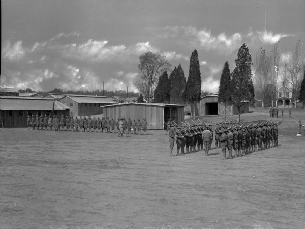 View across grounds toward soldiers during drill practice at Camp Meade, established in 1917. Men are lined up on the left in front of military buildings, and two lines of men holding firearms face each other on the right while an officer gives orders.