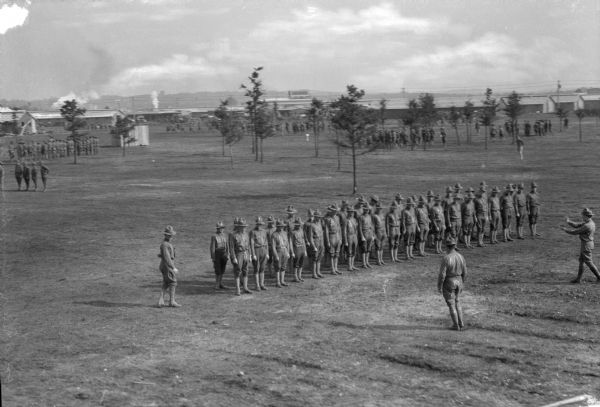 Elevated view of a platoon of men in two rows and their officers on a drill field at Camp Devens, built in 1917. Other soldiers and military buildings are visible in the background.