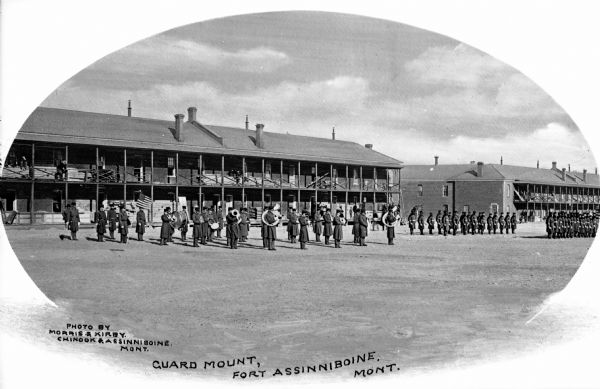 A military band stands in formation in front of a military building at Fort Assinniboine, built in 1879. The United States flag stands near the band and a group of soldiers stands on the right. Caption reads: "Guard Mount, Fort Assinniboine, Mont."