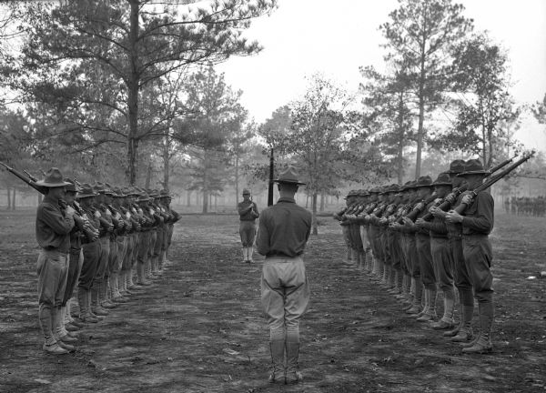 Two rows of soldiers holding firearms stand facing each other in a wooded area at Camp Logan. An officer stands on each side, holding his own firearm, ready to give orders.