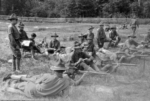 A view of a group of men during rifle practice.  A fence divides the practice field from the wooded background.