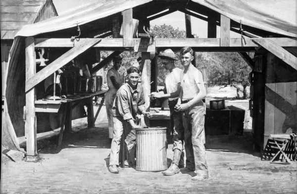 A view of men preparing and serving food from the kitchen tent.  A sign above the group of men reads, "Keep out of kitchen and don't use kitchen utensils."