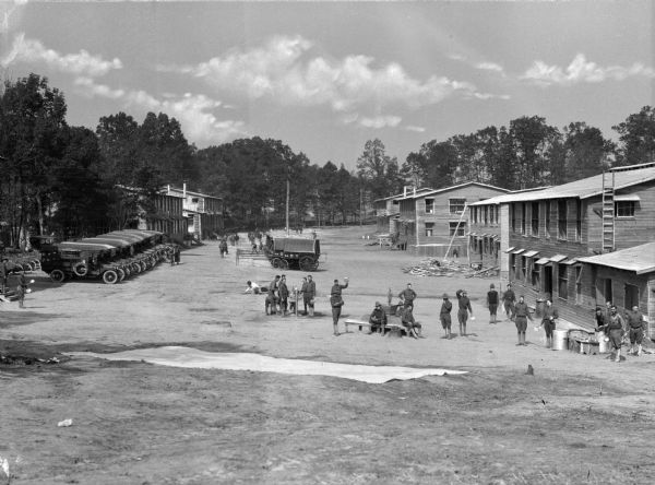 A view of soldiers washing their mess kits outside military buildings, found on the right.  A line of automobiles are parked on the left.