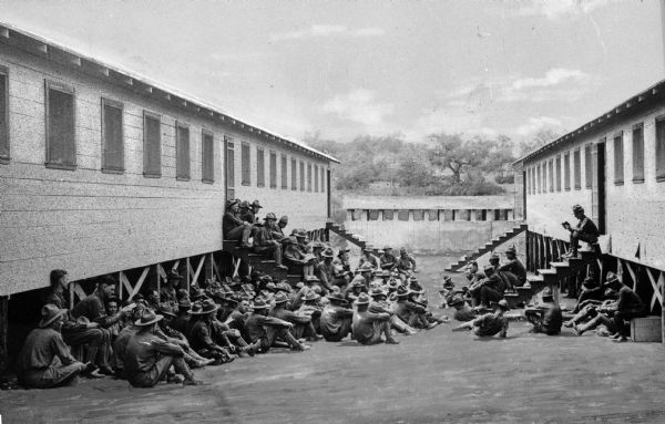 A view of soldiers sitting on the ground and on the steps of military buildings, reading the mail at Camp Travis, named in 1917.