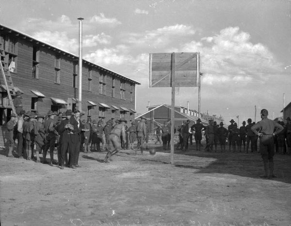 Soldiers standing outside a military building at Camp Upton, built in 1917.  Some soldiers play basketball while others observe.
