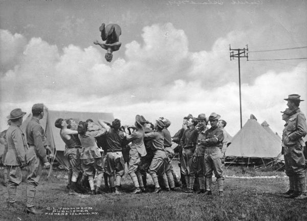 A circle of military men throw a man high up into the air while others look on. Military camp tents are in the background.