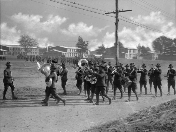 A view of the 316th Regimental Band marching down a dirt road at Camp Meade, established in 1917, with soldiers training and military buildings in the background.