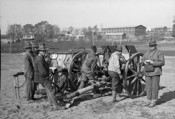 A view of seven soldiers practicing with long distance firing equipment in a field at Camp Meade, established in 1917.  Camp buildings are visible in the background.