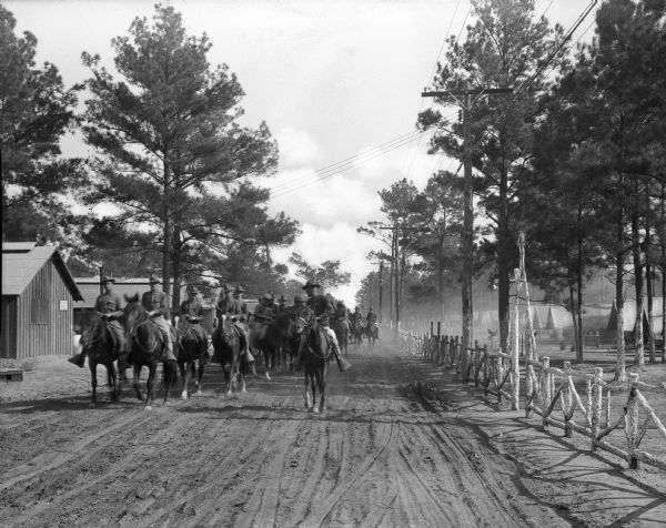 A view of soldiers on horseback, riding down 29th Street at Camp Logan, established in 1917. A fence lines the dirt road on the right, and military buildings stand on the left.