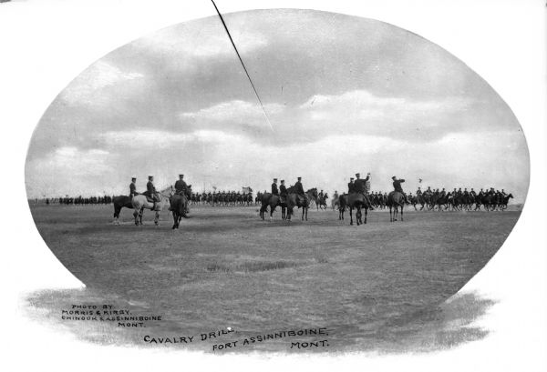 A view of soldiers on horseback leading a cavalry drill. Flags are visible in the formation of men at Fort Assinniboine, built in 1879. Caption reads: "Cavalry Drill, Fort Assinniboine, MONT."