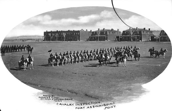 A view of a group of men on horseback during a cavalry inspection at Fort Assinniboine, built in 1879. One soldier holds a flag, and behind the formation stand several military buildings. Caption reads: "Cavalry Inspection, Fort Assinniboine, MONT."