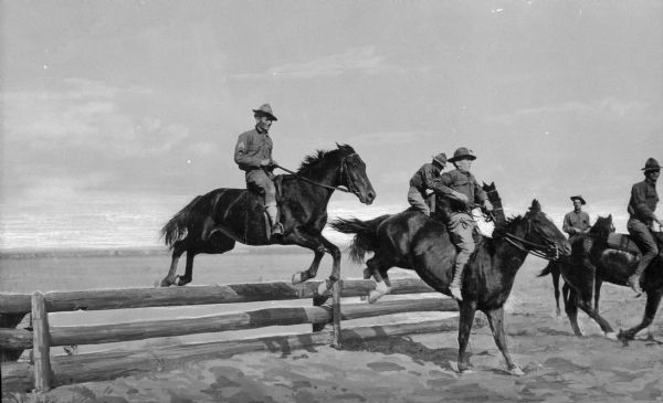 A view of five men on horseback during a cavalry exercise. The horses are jumping over wooden fences on the plain at Camp Travis, named in 1917.