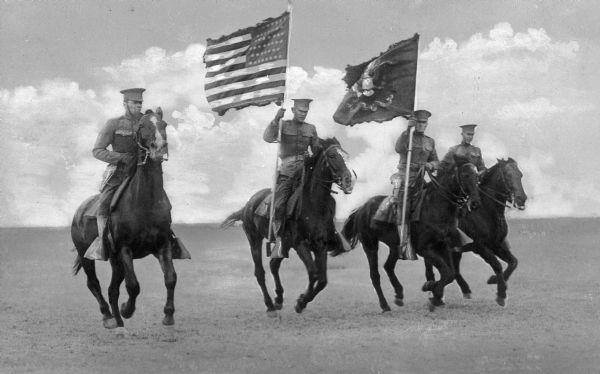 A view of four uniformed men on horseback, two of which hold flags. The soldiers are seen riding across a plain at Camp Travis, named in 1917.