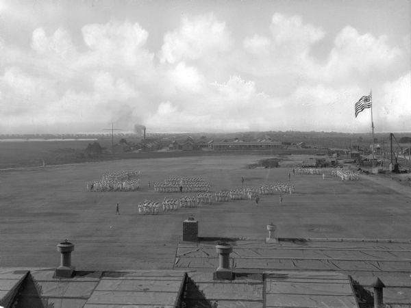 Elevated view of the parade grounds at United States Navy Camp Wissahickon.  Men stand in formation on the parade grounds, and to the the right, a flag waves on a pole.