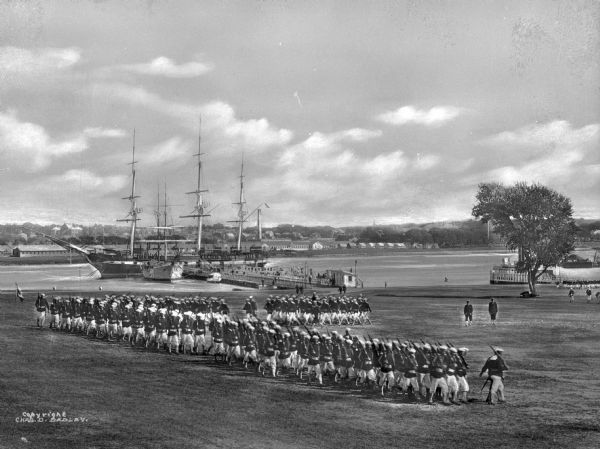 A view of sailors marching on the South Drill Field at the Naval Station Newport, established in 1883. In the background, a warship is in the Narragansett Bay.