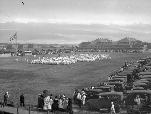A view of a formation of sailors during an exhibition drill at Naval Station Newport, established in 1883. A United States flag and military buildings stand in the background while spectators and their parked automobiles are on the right.