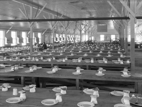 View of tables set in the mess hall at United States Navy Camp Wissahickon.  Two men sit at a table on the left, and windows line the walls.