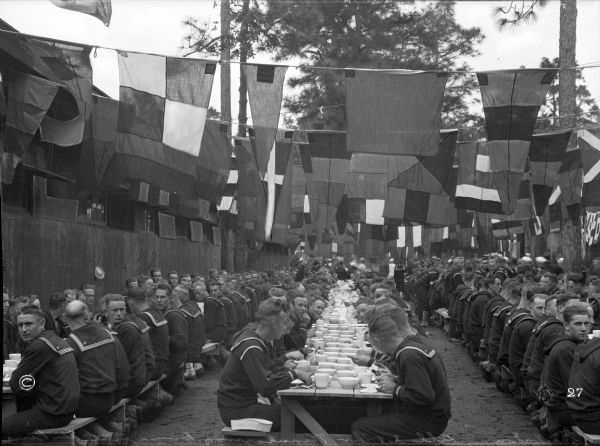 A view of sailors eating at long tables under the pines at a U.S. Naval Training Camp.  Decorative flags wave above them.