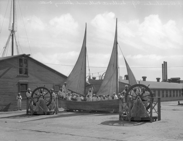 A group of sailors sit in a sailboat while others steer during a sailboat and steering drill at the United States Navy Camp Wissahickon.  Others observe the drill, one out of the window of a military building in the background.