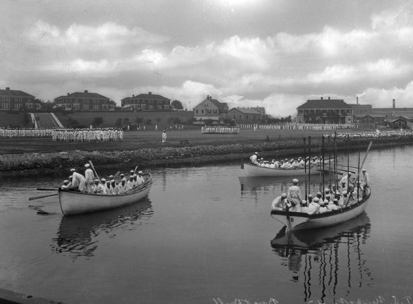 A view of three boats during a boat drill at the Naval Station Newport, established in 1883. They row past the shore on which other formations of sailors execute land drills. Camp buildings stand on the hill behind them.