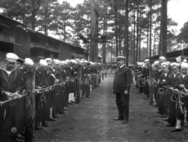 A view of two lines of sailors practicing tying knots in a wooded area at the United States Naval Training Camp.  An officer stands in the center on a dirt path between military buildings.