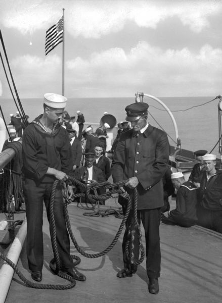 A view of a sailor and an officer tying a reef knot on a United States Merchant Marine Training  Ship.  Other sailors observe and behind them, a flag waves at the front of the ship.