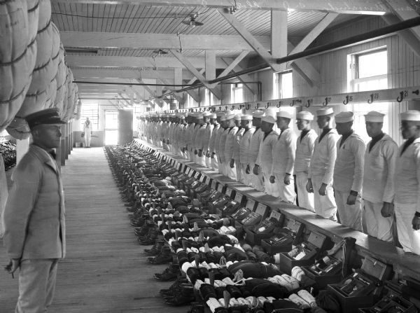 A row of sailors stand in the barracks by their belongings for a clothing inspection.  An officer stands on the left.