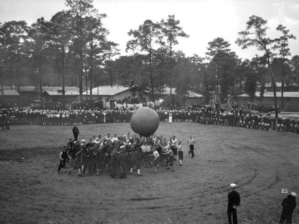 A group of soldiers play with a large push ball while others look on.  The military buildings of the United States Naval Training Camp stand behind the spectators.
