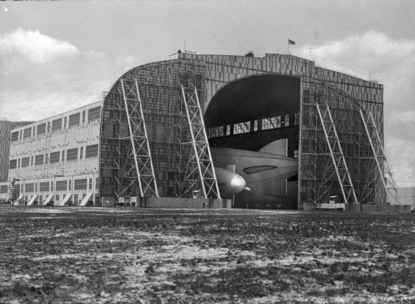 A view of the exterior of Hangar No. One of the Naval Air Station.  A flag stands atop the steel structure, and through the open section, an airship can be seen inside the hangar, erected in 1920.