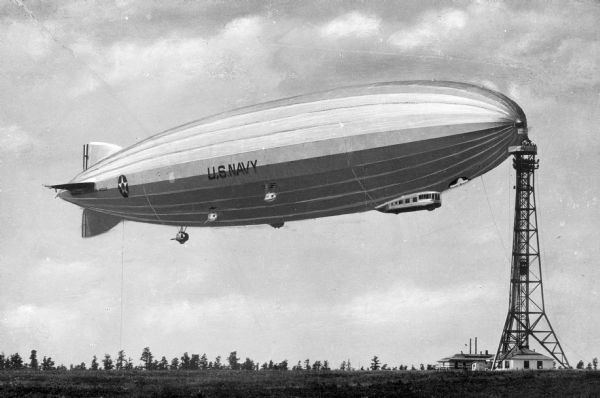 A view of the "Los Angeles" refueling at the Naval Air Station. The airship, completed in 1924, is seen at a tower, flying over a field.
