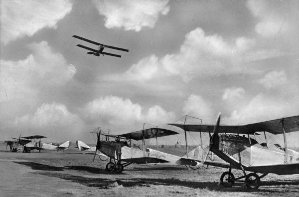 A view of early twentieth century United States Army airplanes on an air field at Camp Vail, named in 1909.  One plane flies overhead, and a man is seen standing on the tire of a grounded plane.