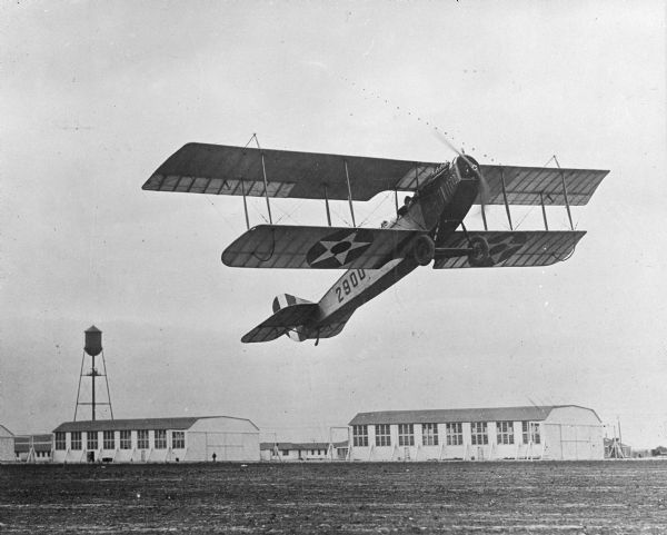 A view of a plane marked with the number 2900 taking off from Kelly Field, created in 1916.  A water tower and the military buidings of Camp Travis are seen in the background.