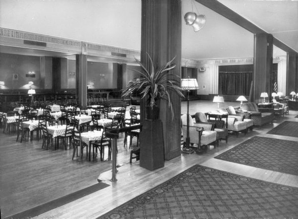 A view of the ballroom of the operating Jewish Agency Board of the United Service Organization Club, a nonprofit organization that provides morale and recreational services to members of the United States Military, established in 1941. Tables are set on the left side of the ballroom, and lounge chairs and tables with lamps are found on the right, where decorative rugs lie on the hardwood floor. The back wall exhibits a stage with curtains.