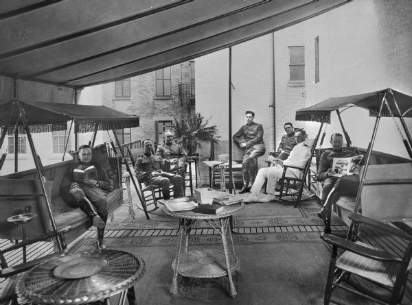 A view of seven men reading and relaxing on the rooftop garden of the Central Park Officers' House. The men sit on outdoor furniture, and behind them, building exteriors are visible.