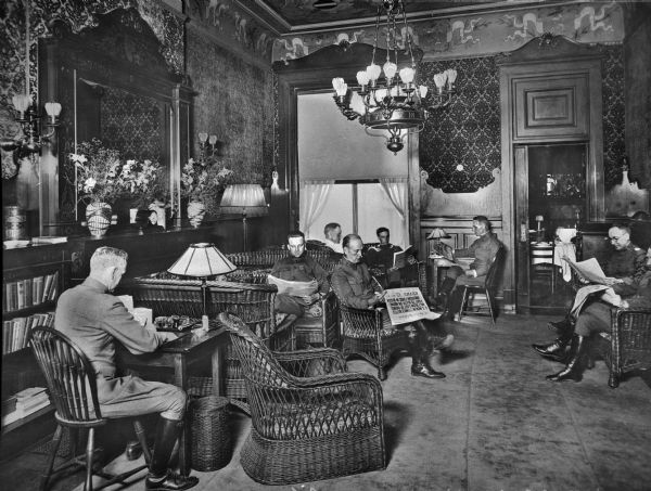 An interior view of the library of the Central Park Officers' House.  Seven men read in wooden and wicker chairs and decorative lighting hangs from the ceiling and on the walls.