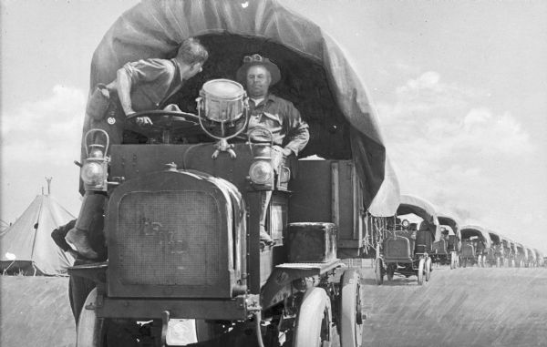 A view of a motor truck train at Camp Travis, named in 1917. The driver sits in the first truck while another man stands beside him.