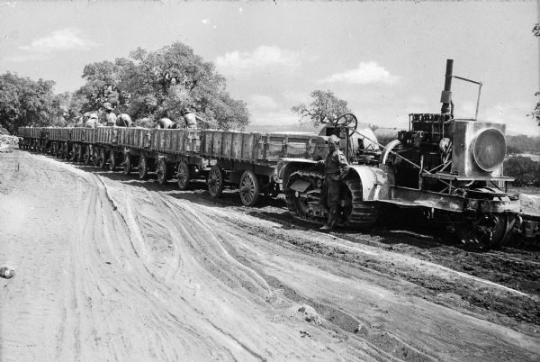 A man stands in front of a Holt Caterpillar tractor at Camp Travis, named in 1917. The tractor pulls a train of wagons on a dirt path. Several men work in the wagons while the tractor is parked.