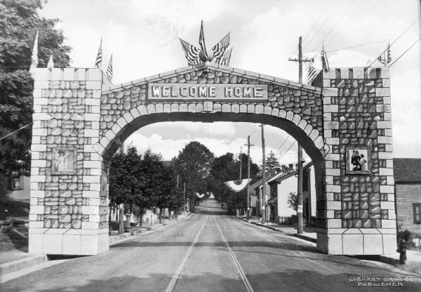 A view down a road with an arch that reads, "Welcome Home." Atop the stone arch stand three flags, and two pictures are on each side. Flags are seen on the side of the road with telephone poles and houses.