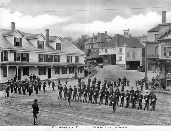 Elevated view of the assembled military men of Company E and the military band on a town street. Other people stand and observe behind the formation of soldiers. On the left stands the Putnam House, and in the distance is a livery store, stable, and dwellings. A horse-drawn carriage is parked on the right. Caption reads: "Company E. Orange, Mass."