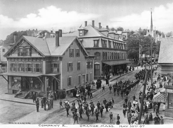 Elevated view of a military parade marching through a town street. A band leads the soldiers of Company E, and spectators can be seen on either side of the street, which is lined with stores, churches, and other buildings. Caption reads: "Company E. Orange, Mass. May 30th 07."
