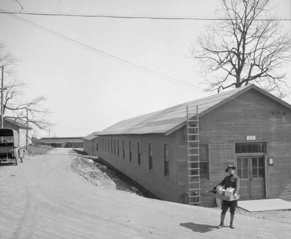 Exterior of the United States Army General Hospital, Building 23. A uniformed man stands outside, carrying a tray. A dirt road leads to the left where an automobile is parked.