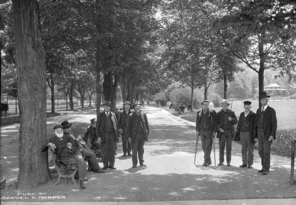 A group of men posing on a parkside path. Some of the men occupy benches, while others stand on the path.